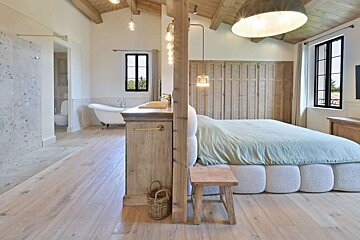 Spacious, bright bedroom with an open-plan bathroom featuring a clawfoot tub, modern bed, and light wood finishes. Vaulted ceiling and large windows.