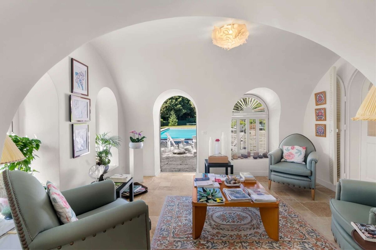 An elegant white arched living room with light green armchairs, a coffee table, and a patterned rug, looking out onto a bright blue swimming pool.