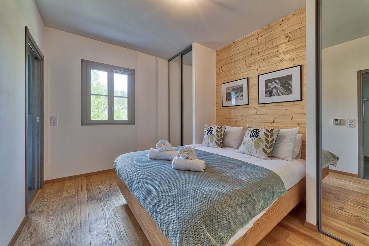 A cozy bedroom with a wooden bed, accent wall, and floor. It features a window, sliding mirrored closet, and a bed with decorative pillows and towels.