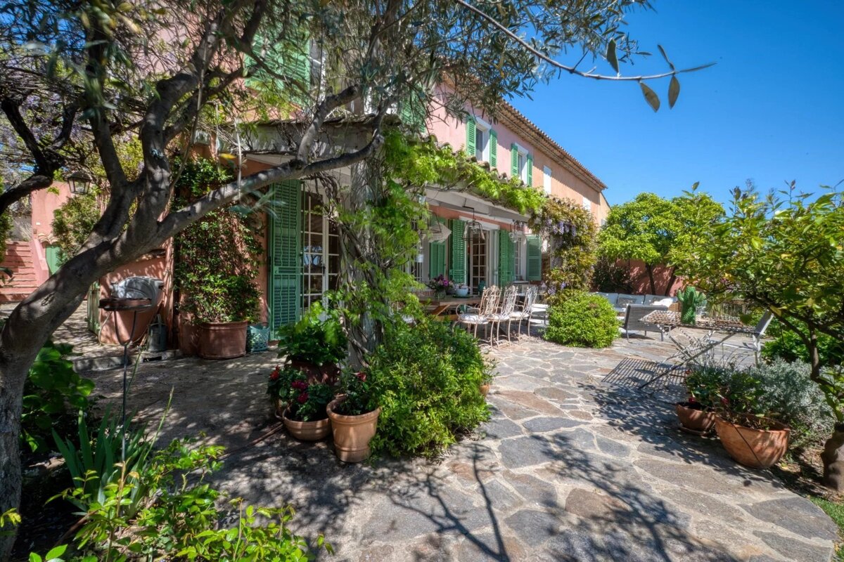 Sunny Mediterranean villa with peach walls, green shutters, a stone patio, and vibrant greenery under a clear blue sky.