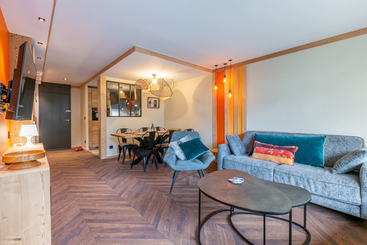 A modern open-plan living/dining room with warm orange accents, chevron wood floors, a gray sofa, and a sleek dining set, radiating modern warmth.