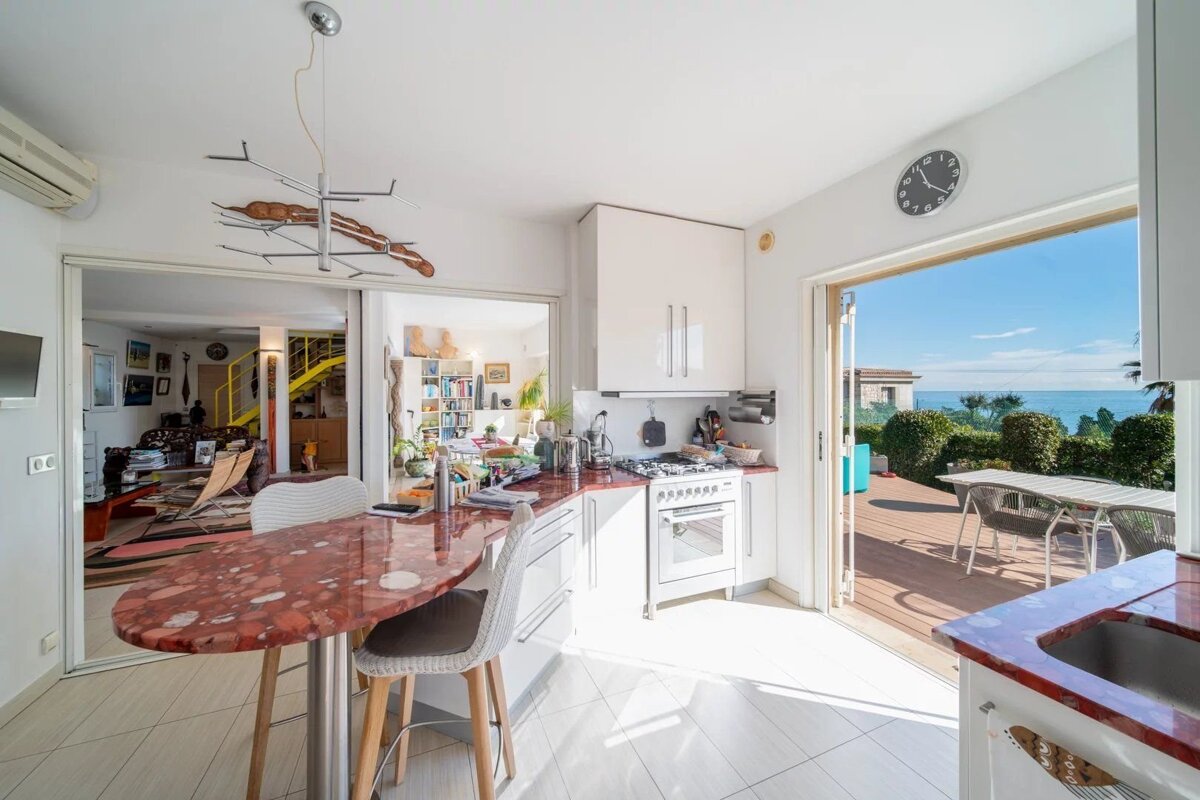 Bright kitchen with red marble counters, bar, and white appliances, opening to a sunlit deck with a stunning ocean view. Living room visible through glass doors.