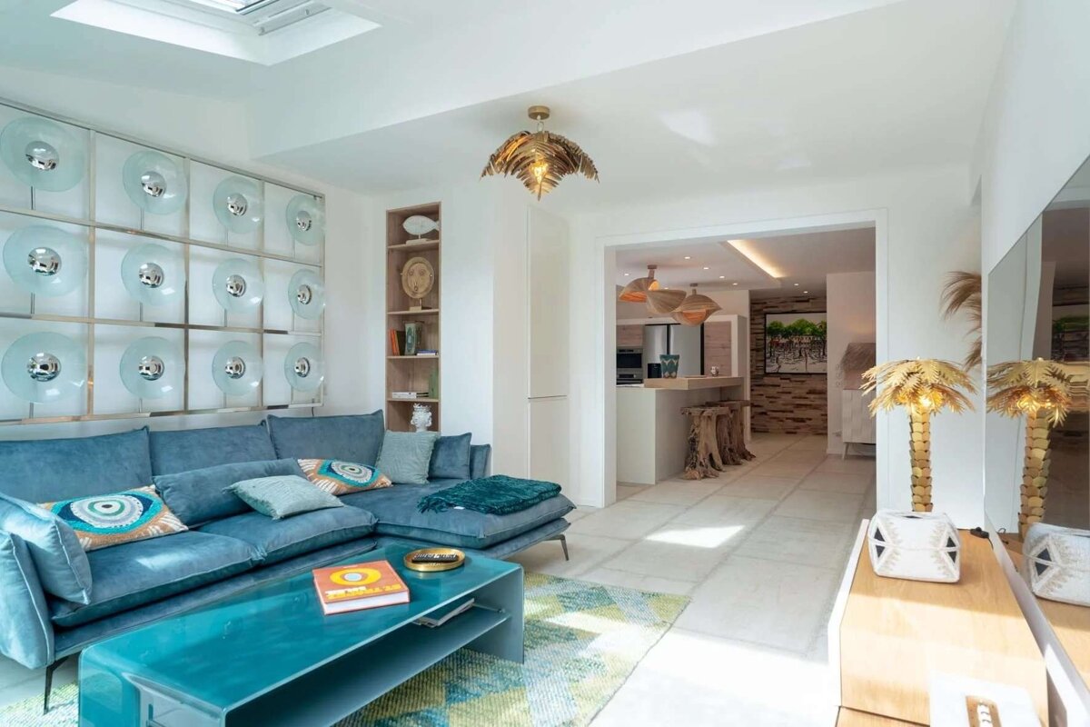 A vibrant living room features a blue sectional, teal coffee table, and striking disc wall art. It opens to a modern kitchen with unique bar stools.