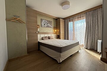 A cozy, modern bedroom with wooden floors, a large bed with a white headboard, wood paneled walls, and sheer curtains over a balcony window.