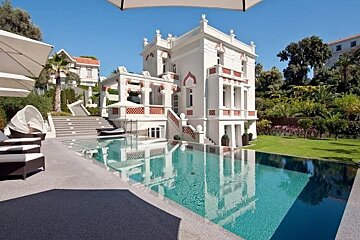 Luxurious white villa with Moorish architectural details and a large, clear swimming pool reflecting the building.