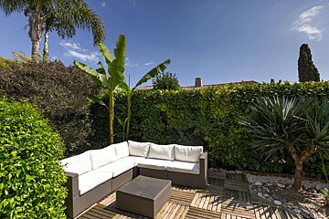 Sunny outdoor patio with a rattan sectional sofa, coffee table, and lush greenery including a large hedge, palm, and banana trees.