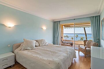 A serene bedroom with light blue walls, a large bed, and direct access to a balcony boasting stunning ocean views and palm trees.