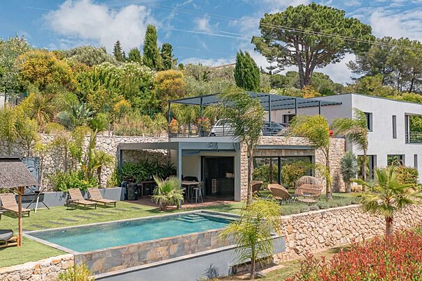 A modern luxury house with a swimming pool and lush terraced gardens on a sunny hillside, featuring a car parked on an upper level.
