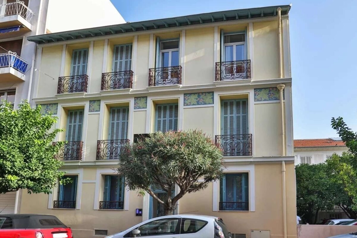A light yellow building with blue shutters, ornate wrought iron balconies, and decorative green friezes. Trees and parked cars are in front.