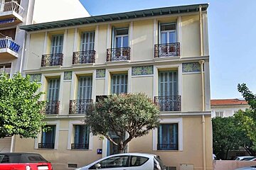 A light yellow building with blue shutters, ornate wrought iron balconies, and decorative green friezes. Trees and parked cars are in front.