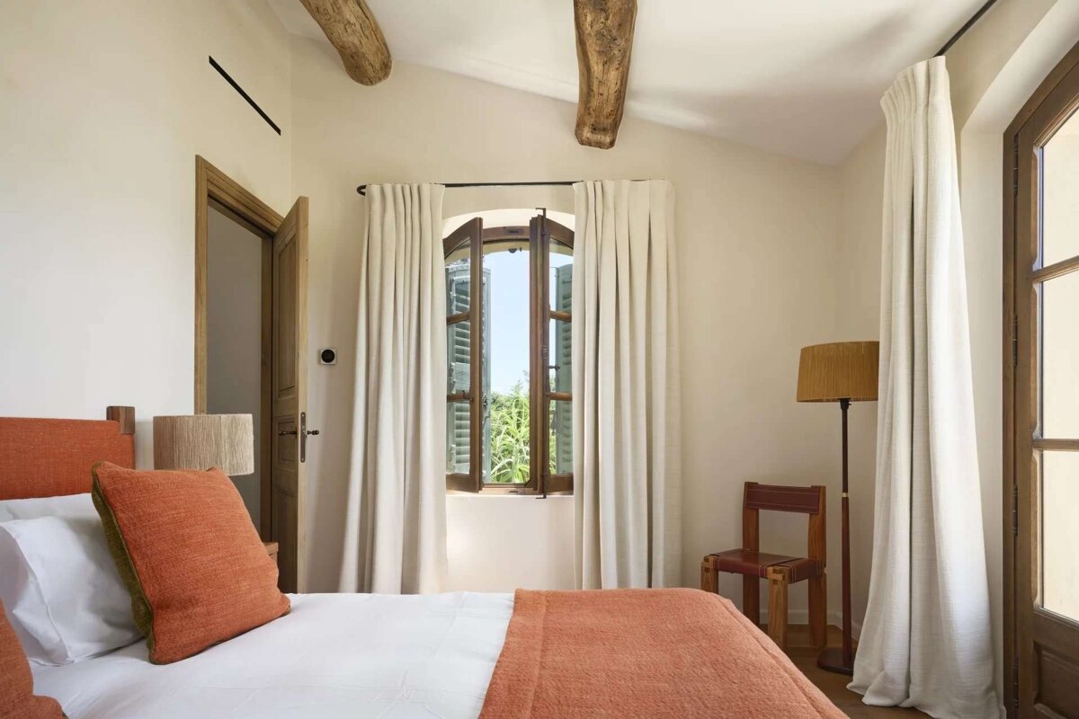 A bright, rustic bedroom with white walls, exposed wooden beams, and a bed with orange linens. A window with open shutters looks out to greenery.