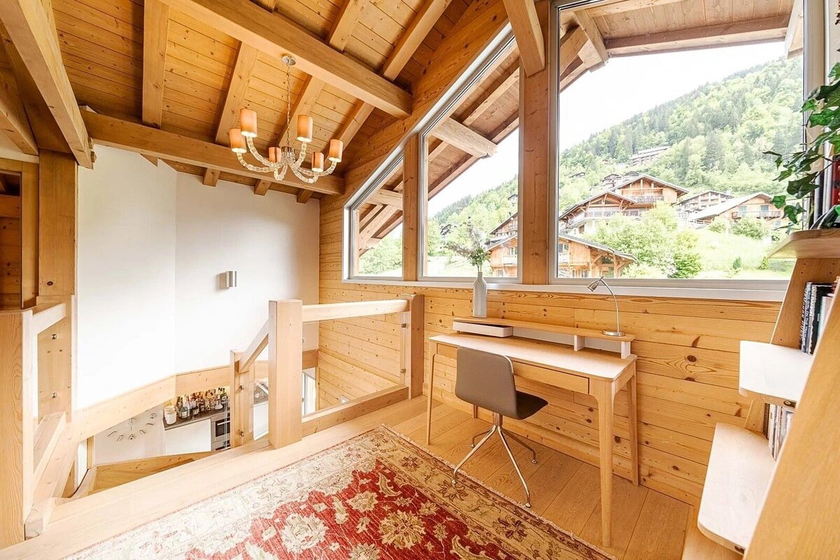 Bright, wood-paneled loft office with a desk, large windows revealing a scenic mountain village, cozy rug, and chandelier.