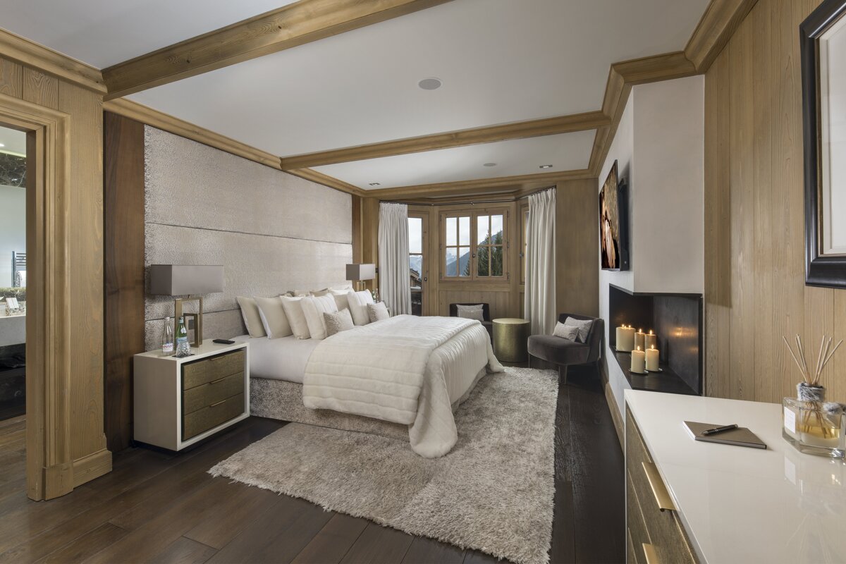 Luxurious chalet-style bedroom with warm wood paneling, a plush white bed, a shag rug, and a cozy fireplace with candles.