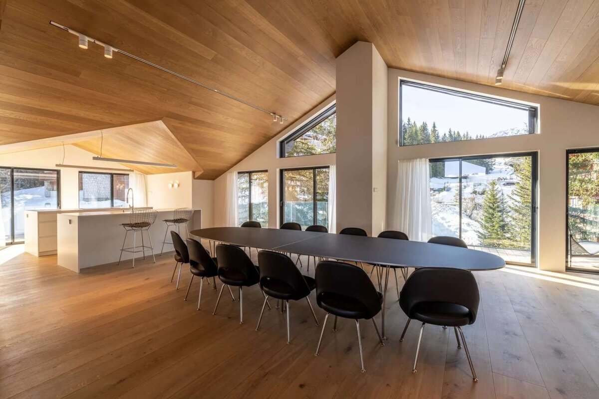 Spacious, modern dining room & kitchen with wooden vaulted ceilings, large windows revealing a snowy mountain and forest view, and a long black table.