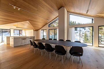 Spacious, modern dining room & kitchen with wooden vaulted ceilings, large windows revealing a snowy mountain and forest view, and a long black table.