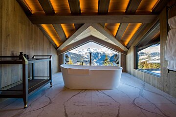 A luxurious attic bathroom with a freestanding tub offers breathtaking panoramic views of snow-capped mountains and pine trees.