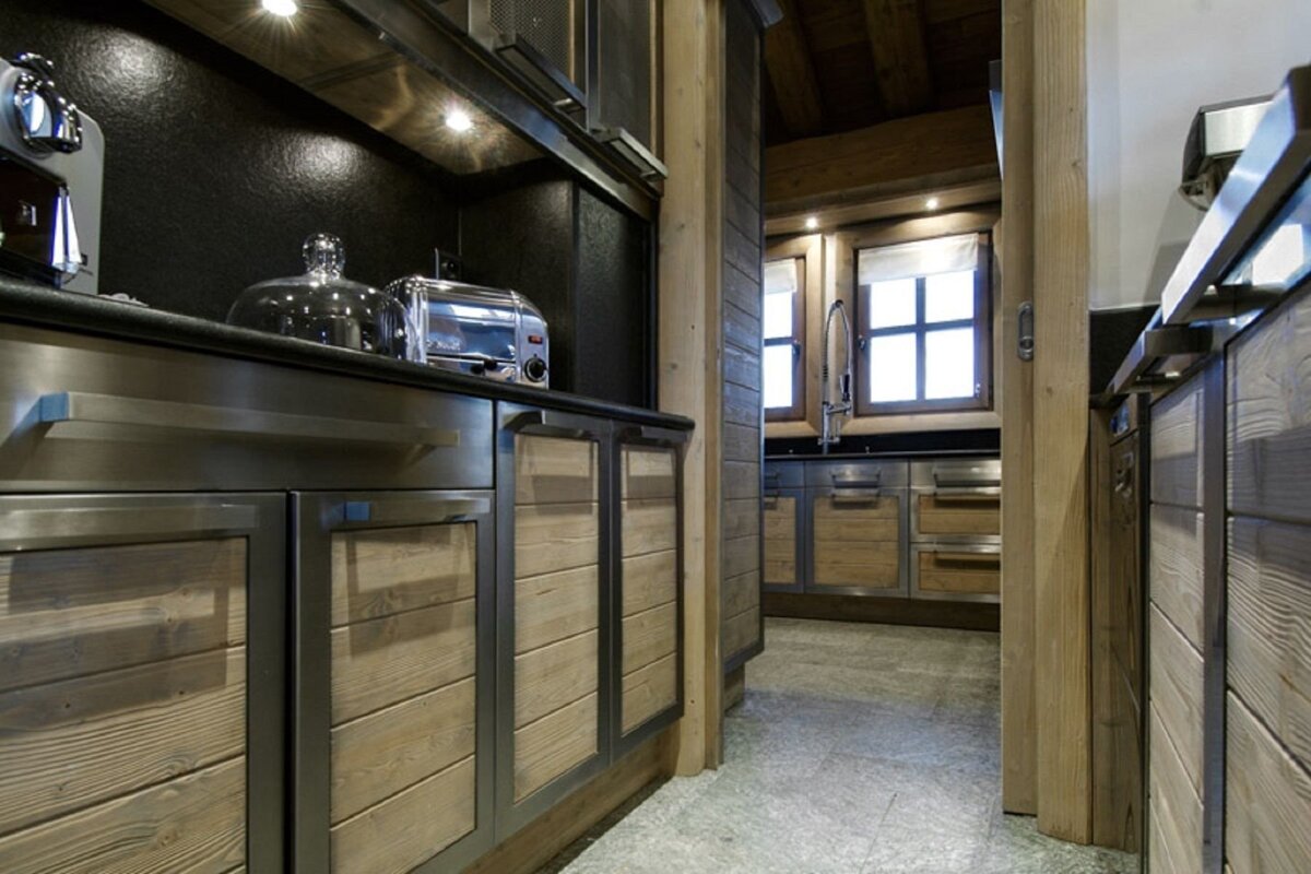 A cozy, rustic-modern kitchen features light wood and stainless steel cabinets. A toaster is on the counter, with a sink and windows visible in the back.