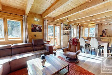 Cozy, sunlit alpine chalet interior with wooden walls, a large brown sofa, coffee table, armchair, and dining area.