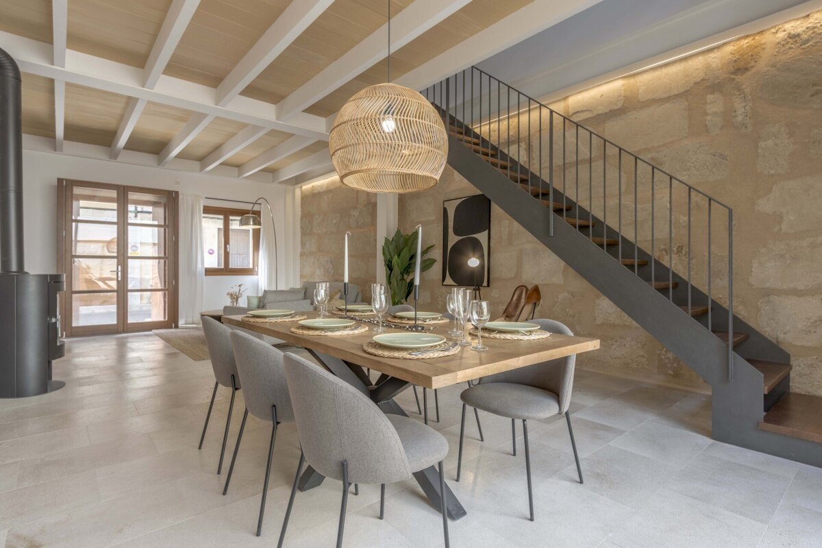 A modern dining room features a wooden table, grey chairs, wicker pendant light, exposed stone wall, and a sleek metal staircase.
