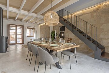 A modern dining room features a wooden table, grey chairs, wicker pendant light, exposed stone wall, and a sleek metal staircase.