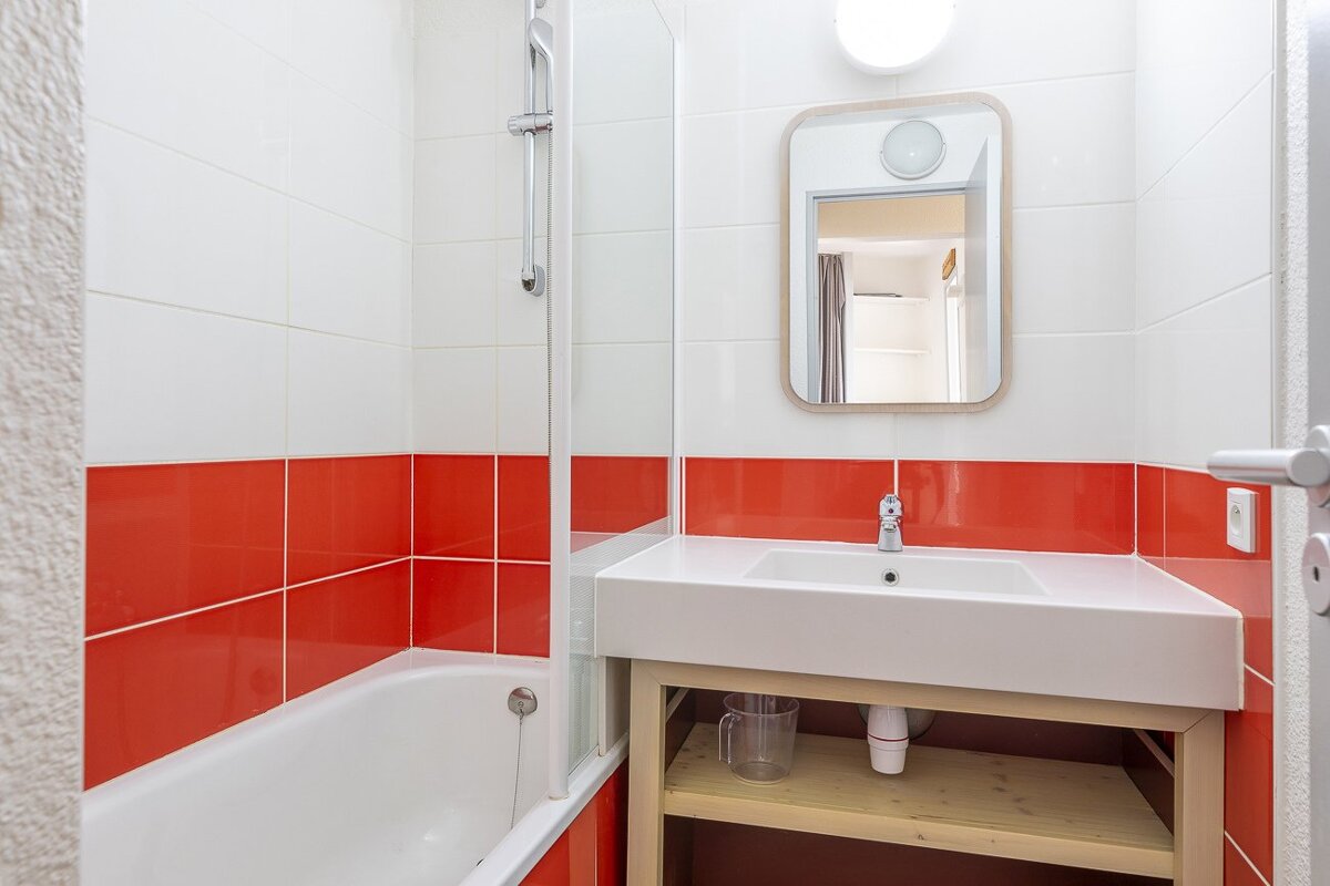 A bright bathroom with white and red tiled walls, a bathtub with shower, a vanity sink, and a rectangular mirror above it.