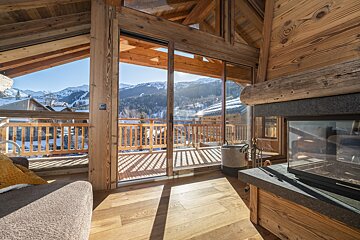 Sunlit wooden chalet living room with a fireplace and large glass doors opening to a balcony, offering stunning views of snowy mountains and ski slopes.