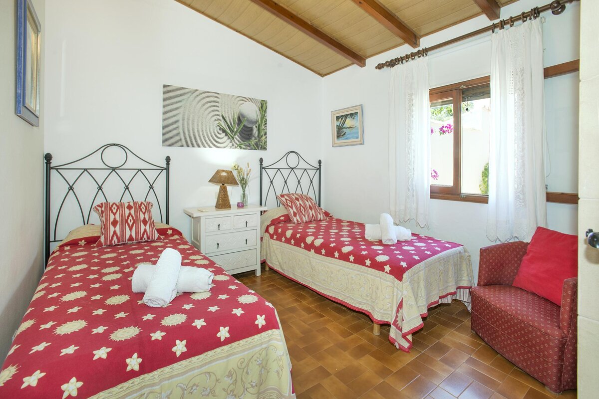 A bright bedroom features two twin beds with red and white bedding, a tiled floor, and a wooden beamed ceiling. A window with white curtains adds light.