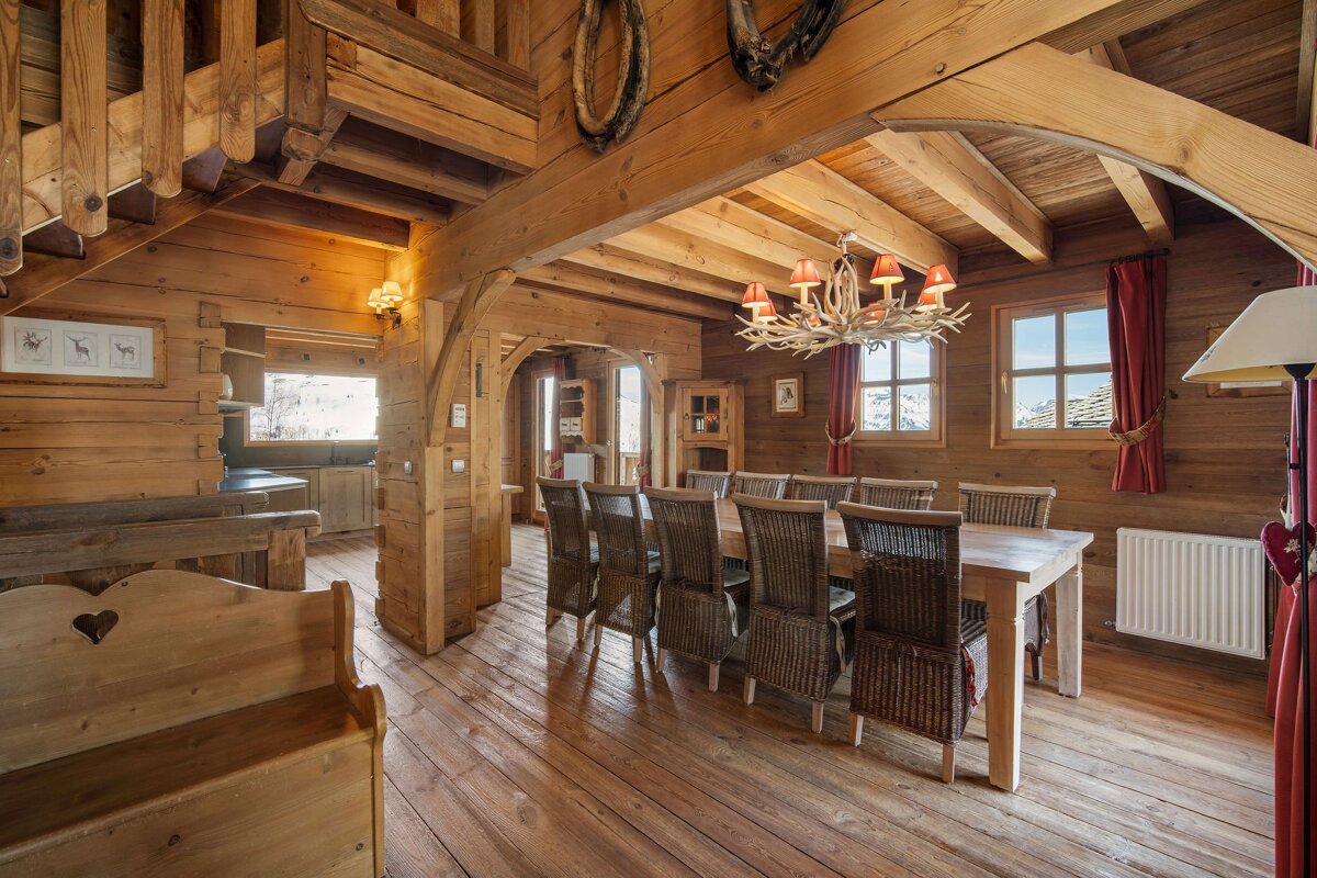 A cozy, rustic wooden cabin interior featuring a long dining table with wicker chairs and a unique antler chandelier.