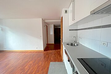 An empty room with warm wood flooring, white walls, and a modern kitchenette on the right featuring a sink, stovetop, and white cabinets.
