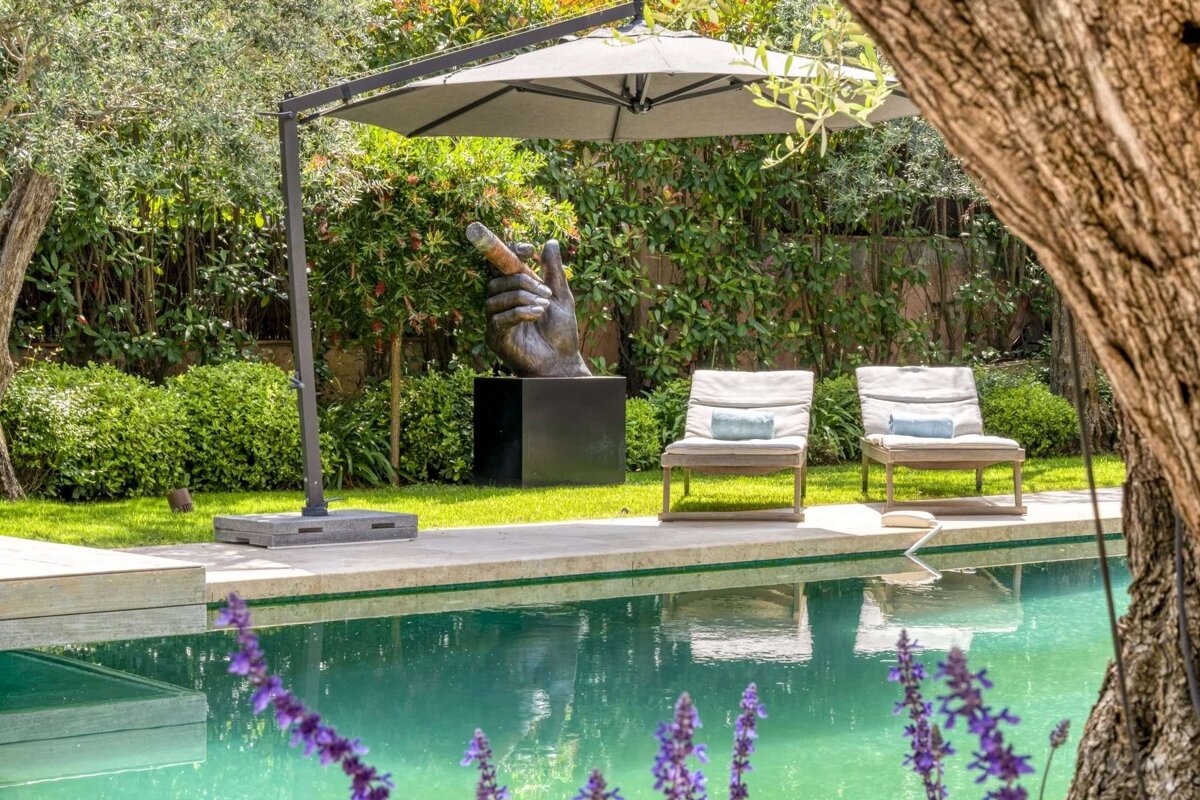 A luxurious poolside scene featuring a hand sculpture holding a cigar, lounge chairs, an umbrella, and lush greenery, with purple flowers in the foreground.