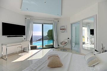 Sleek white bedroom features a TV, desk, and a stunning ocean view with an infinity pool visible through large glass doors.