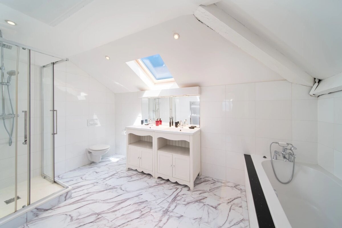 Bright white bathroom featuring marble flooring, a glass shower, a double vanity, a bathtub, and a skylight in the slanted ceiling.