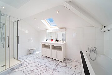 Bright white bathroom featuring marble flooring, a glass shower, a double vanity, a bathtub, and a skylight in the slanted ceiling.