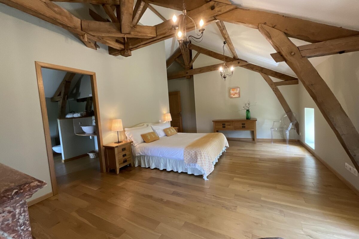 A bright, rustic bedroom features exposed wooden beams, hardwood floors, a large bed with side tables, a dresser, and an open doorway to an ensuite.