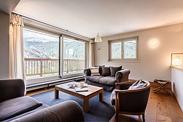 A cozy living room with large windows overlooking mountains and forest. Features comfortable sofas, a coffee table, and warm lighting.