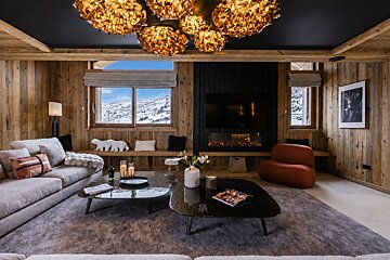 Cozy rustic-modern chalet living room with wood walls, unique gold chandeliers, a fireplace, and a snowy mountain view.
