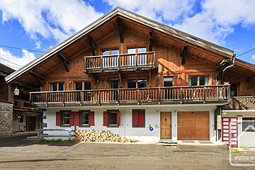 A traditional wooden chalet with balconies, red shutters, a stack of firewood, and a garage, set against a bright blue sky.