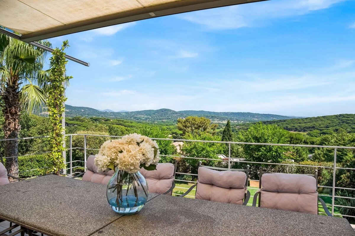 Panoramic view from a sunny terrace with a floral centerpiece, overlooking rolling green hills and mountains under a clear blue sky.