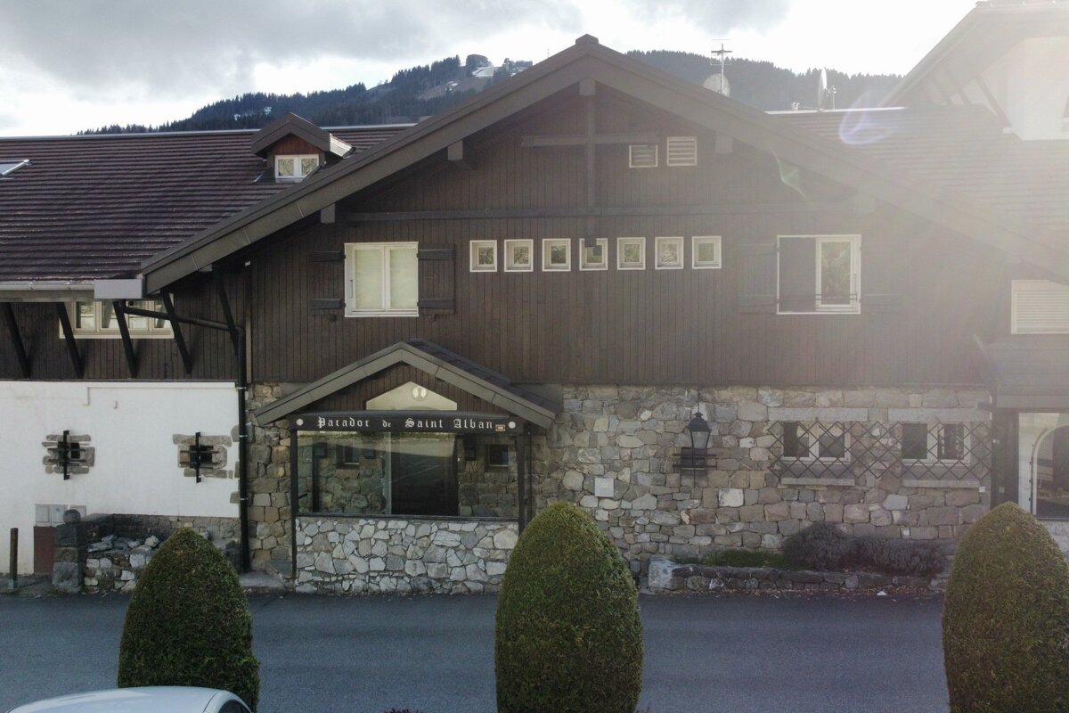 A chalet-style building with a stone lower level, dark wood upper level, and three cone-shaped green bushes in front.