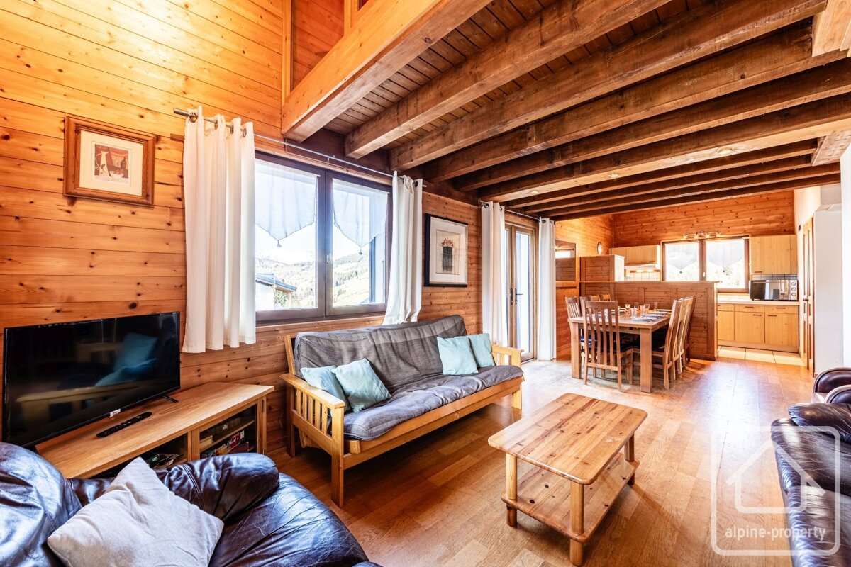 A cozy, rustic wooden chalet interior featuring a living room, dining area, and kitchenette. Exposed beams and mountain views enhance the warm atmosphere.