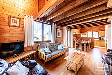 A cozy, rustic wooden chalet interior featuring a living room, dining area, and kitchenette. Exposed beams and mountain views enhance the warm atmosphere.
