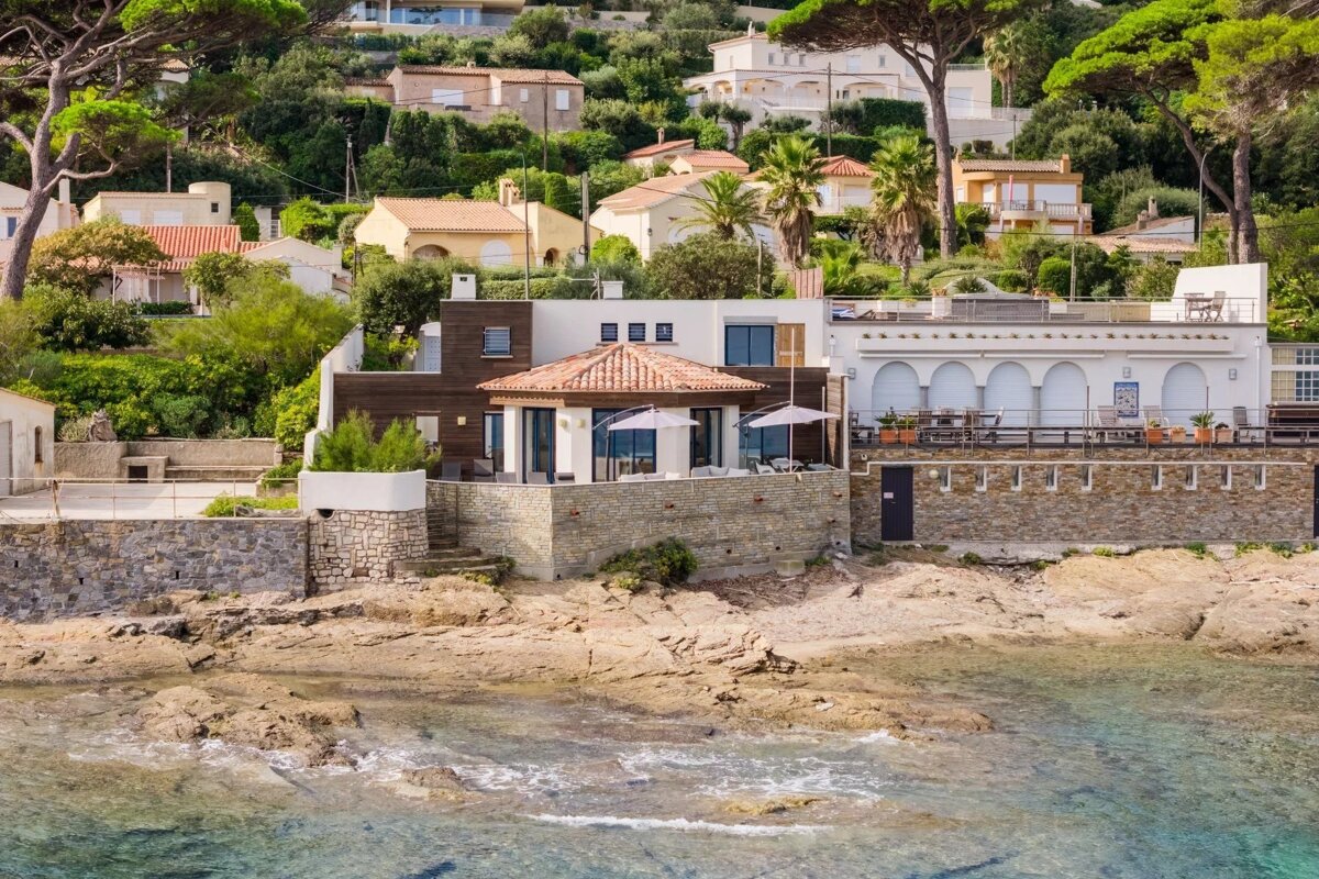 Coastal villas on a rocky shore with clear turquoise water, backed by a lush, green hillside.