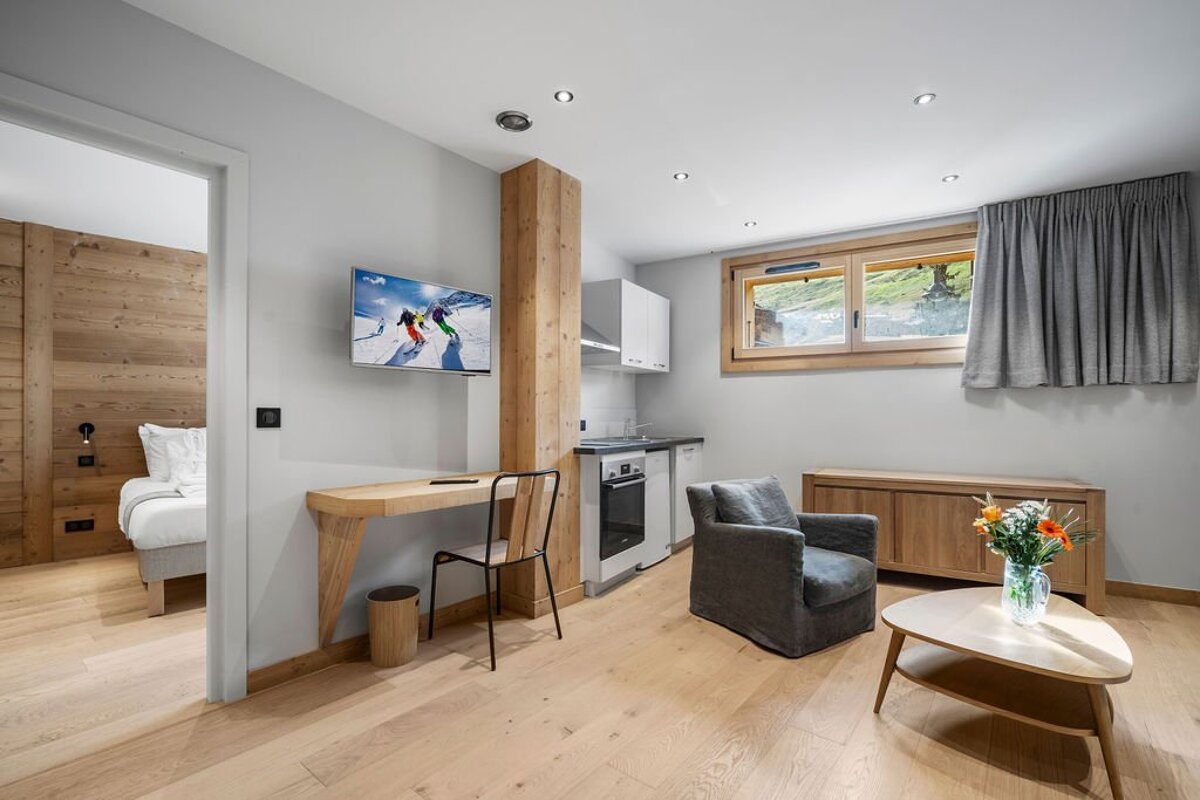 A modern alpine studio featuring a desk with TV, a kitchenette, a cozy armchair, and a doorway showing a wooden bedroom.