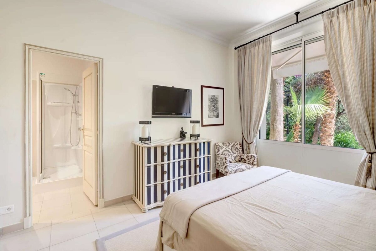 Cream-colored bedroom featuring a bed, striped dresser, TV, large window with palm tree views, and an open door to an en-suite shower.