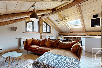 Cozy attic lounge with exposed wooden beams, a brown sectional sofa, and modern light fixtures. Bright windows and a kitchenette complete the space.