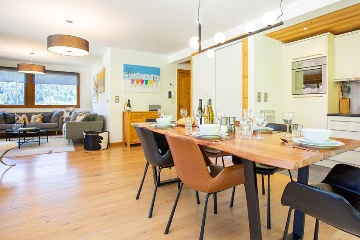 A bright, open-plan home featuring a set wooden dining table with modern chairs, leading to a cozy living area and kitchen.