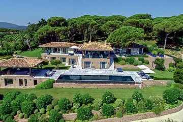 Aerial shot of a sprawling luxurious villa with multiple stone buildings, a long swimming pool, terraced gardens, and a backdrop of pine forests and vineyards.