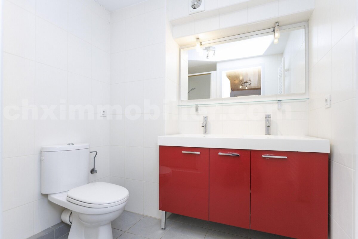 A modern white-tiled bathroom featuring a toilet, a vibrant red double vanity with two sinks, and a large illuminated mirror.