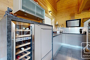 A modern grey kitchen with stainless steel appliances, including a wine fridge, contrasting with rustic wood-paneled walls and ceiling in a cozy chalet.