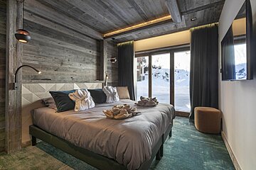 Cozy rustic bedroom with wood-paneled walls, a large bed, and sliding glass doors looking out onto a snowy winter landscape.